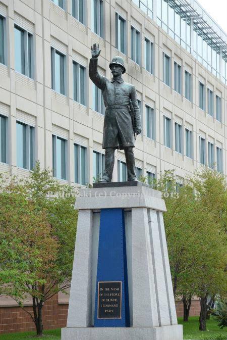 Historiallinen valokuva Chicagon Haymarket-aukiolta. Kuvassa on korkealla jalustalla seisova poliisipatsas, jonka käsi on kohotettu ylös komennukseen. Taustalla näkyy kaupungin korkeita kerrostaloja ja katunäkymää.