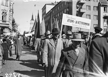 ”Mustavalkoinen kuva vappukulkueesta Oulussa vuonna 1958. Ihmiset kantavat suurta kylttiä, jossa lukee tekstiä atomi- ja vetypommien kieltämisestä.”