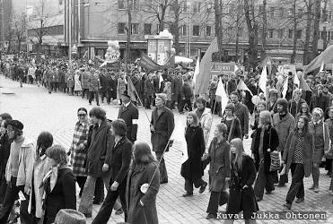 Mustavalkoinen valokuva 1950-luvun Helsingistä. Vappukulkueen osallistujat kiertävät jonossa torin reunaa pitkin, jättäen aukion keskiosan avoimeksi. Joillakin marssijoilla on pieniä kylttejä ja tunnelma on harras.