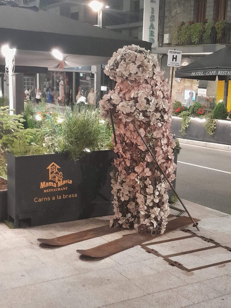 Hiihtävä karhu, joka päällystetty kukilla. Jalassa leveät ruskeat puusukset ja kädessä sauvat. Keskellä kaupungin keskustaa.