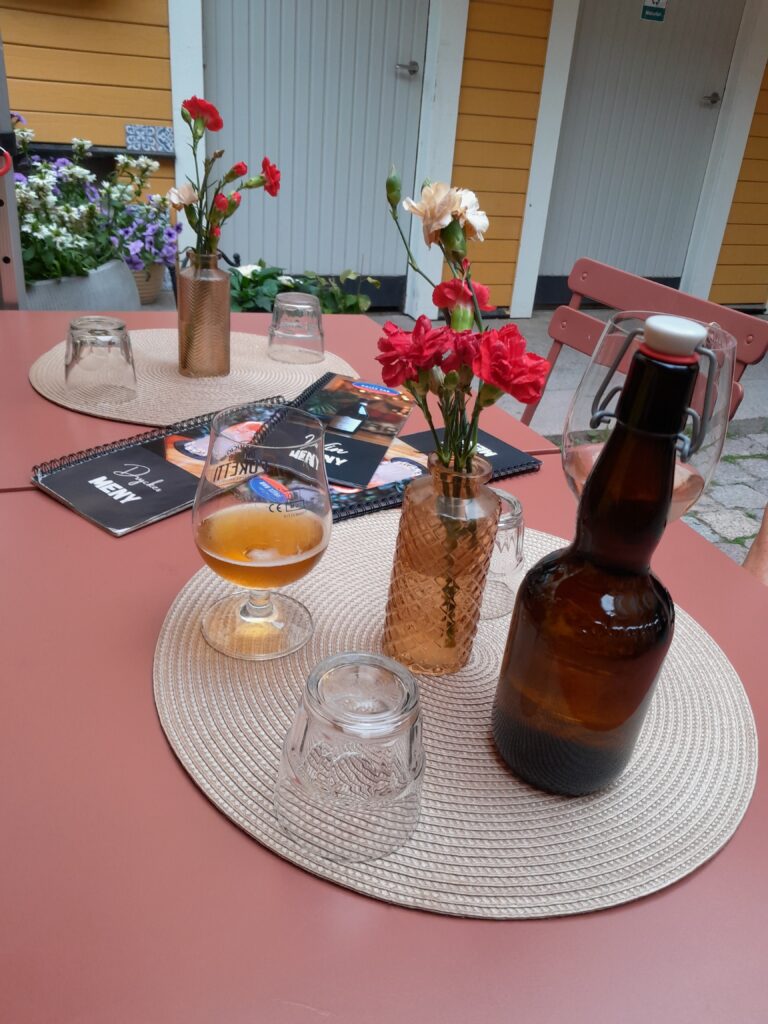 Ravintolan pöydällä "säleinen" pullokukkamaljakko, jossa punaisia neilikoita. Pöydällä myös olutpollo ja -lasi sekä vesilasi.