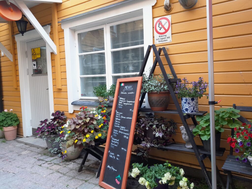 Paljon kukkia kukkahyllyillä ja -telineissä ulkona.  Kuva Nortälljestä ravintolan sisäpihalta.