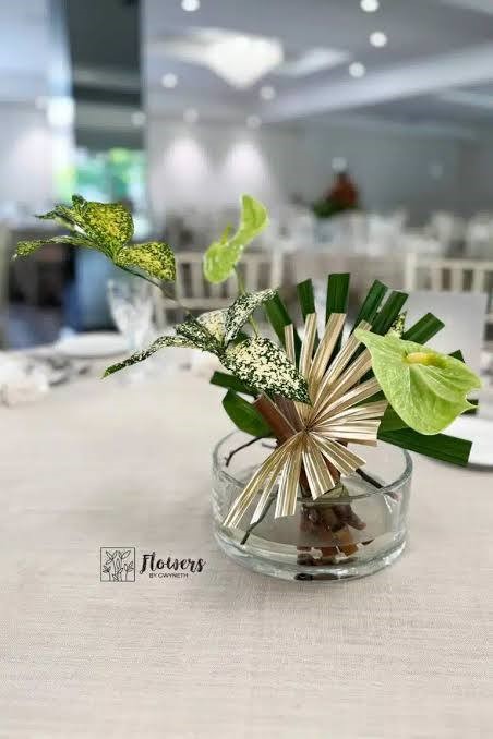 "Matala ja minimalistinen japanilainen ikebana-kukkasetelma pyöreässä, matalassa lasivadissa. Asetelmassa on kaksi flamingonkukkaa, muutama leveä vihreä lehti sekä runsaasti tyhjää tilaa ja se on aseteltu vinoon linjaan."