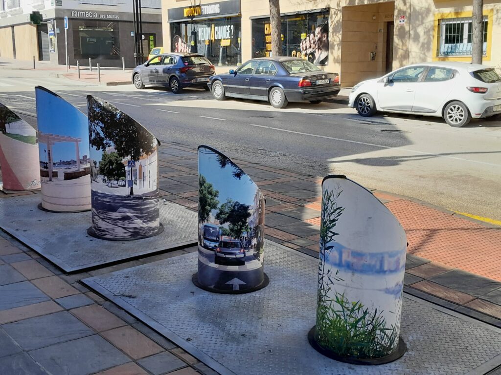 Fuengirola - suomalainen paratiisi Espanjassa on myös katunäkymä , jossa metalliset roskatornit "taiteiltu" maisemakuvilla. Myös autoja parkissa ja liiketiloja näkyy.